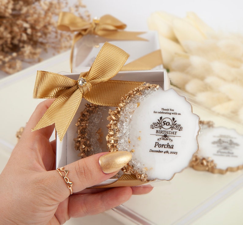 Puede incluir: Una caja de regalo blanca y dorada con una cinta dorada y un regalo redondo blanco y dorado con el texto "Gracias por celebrar con nosotros. 50. Cumplea&ntilde;os Porcha 4 de diciembre de 2023."