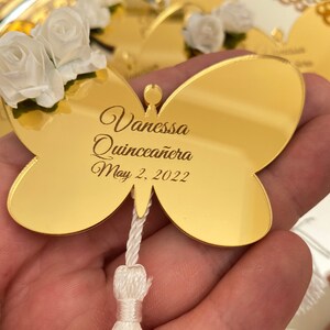May include: Gold butterfly-shaped mirror with a white tassel. The mirror has the text "Vanessa Quincea&ntilde;era May 2, 2022" engraved on it.