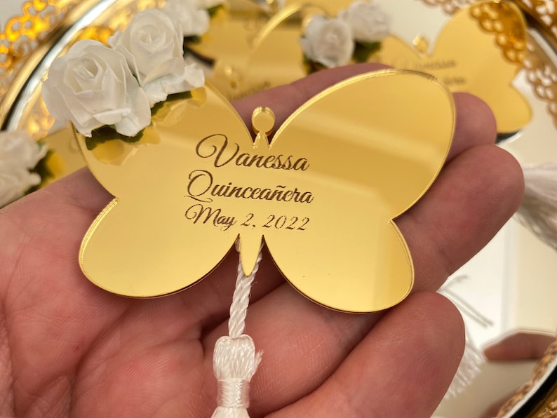 May include: Gold butterfly-shaped mirror with a white tassel. The mirror has the text "Vanessa Quincea&ntilde;era May 2, 2022" engraved on it.