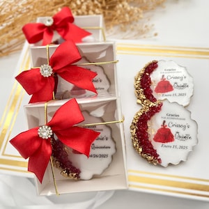 Op de afbeelding: Een set van vijf transparante cadeauboxen met rode strikken en strass. Elke doos bevat een ronde witte koek met een rood en gouden ontwerp en de tekst "Gracias por acompa&ntilde;arme Crissey's Quince Enero 15, 2025".