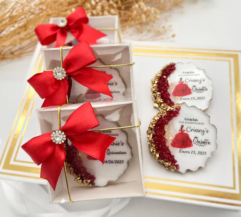 Op de afbeelding: Een set van vijf transparante cadeauboxen met rode strikken en strass. Elke doos bevat een ronde witte koek met een rood en gouden ontwerp en de tekst "Gracias por acompa&ntilde;arme Crissey's Quince Enero 15, 2025".