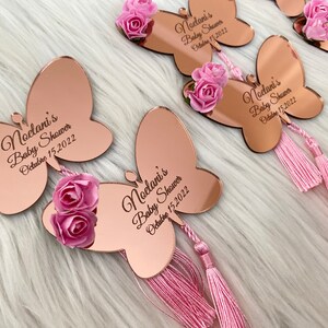 May include: Rose gold mirror butterfly favors with pink flowers and tassels. The butterflies have the text "Noelani's Baby Shower October 15, 2022" engraved on them.