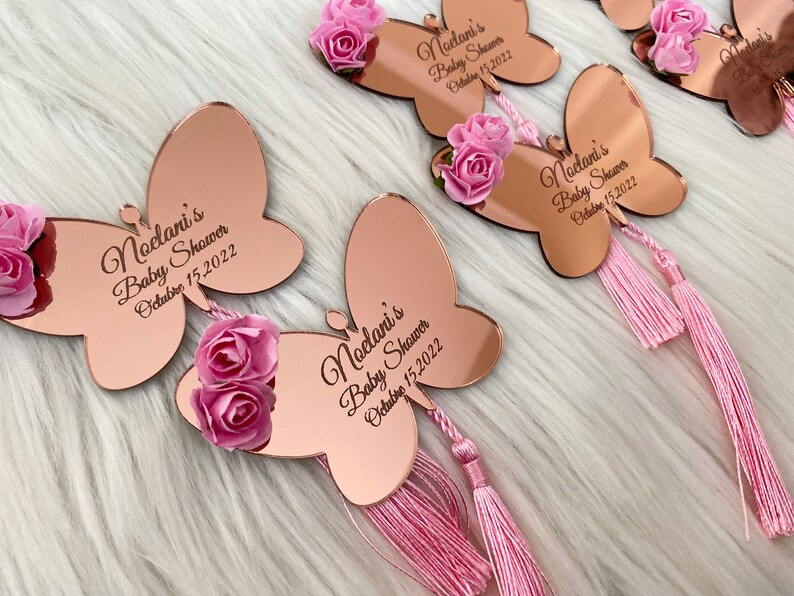 May include: Rose gold mirror butterfly favors with pink flowers and tassels. The butterflies have the text "Noelani's Baby Shower October 15, 2022" engraved on them.