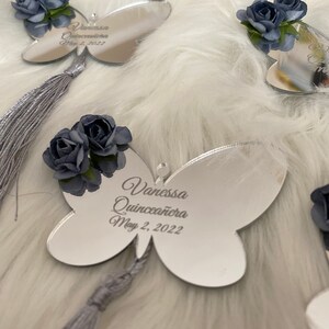 May include: Silver butterfly-shaped mirror with a gray tassel and a blue flower embellishment. The mirror has the text "Vanessa Quincea&ntilde;era May 2, 2022" engraved on it.