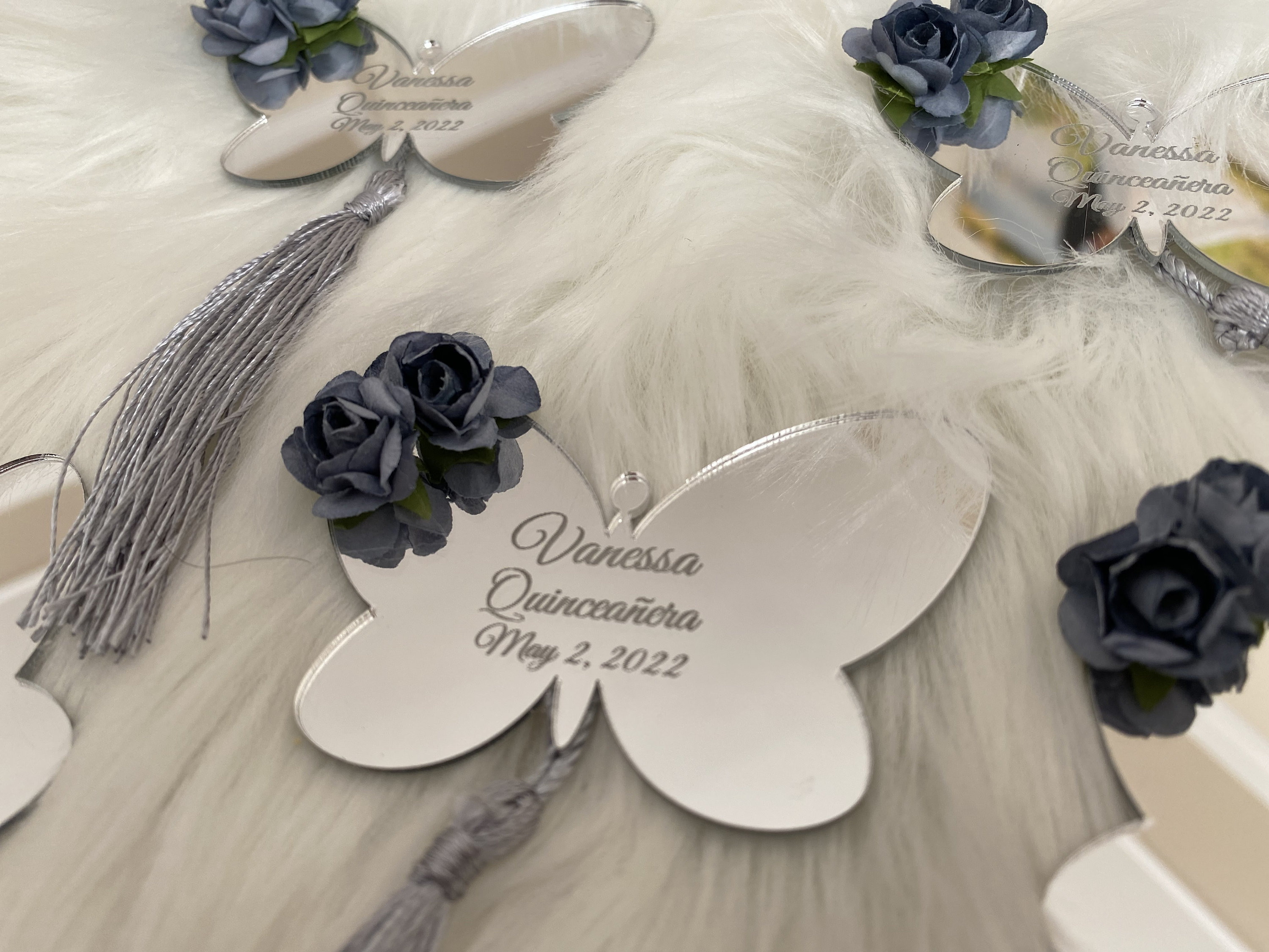 May include: Silver butterfly-shaped mirror with a gray tassel and a blue flower embellishment. The mirror has the text "Vanessa Quincea&ntilde;era May 2, 2022" engraved on it.
