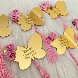 May include: Gold butterfly-shaped acrylic mirror with a pink tassel and a pink flower embellishment. The mirror has the text "Vanessa Quincea&ntilde;era May 2, 2022" engraved on it.