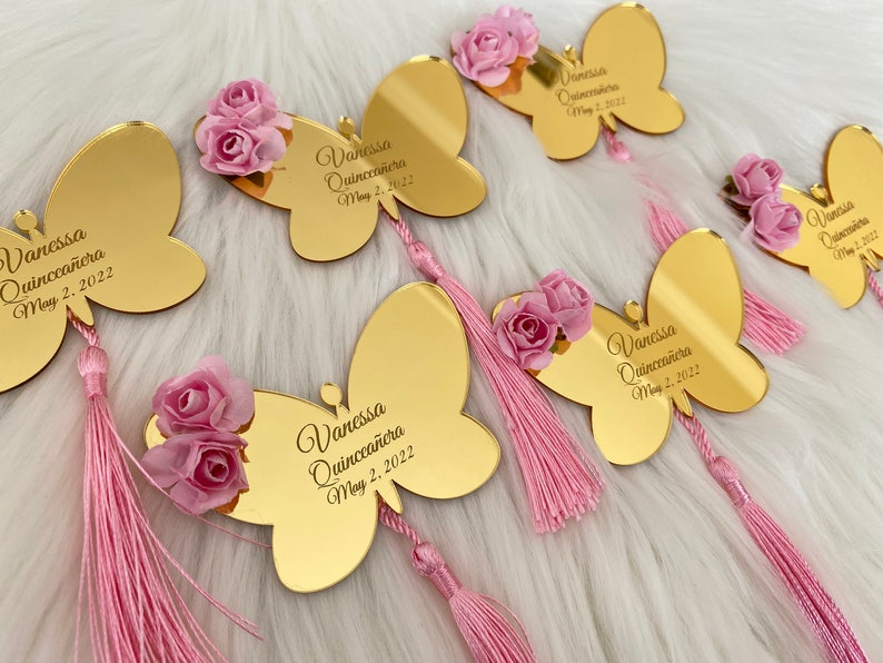 May include: Gold butterfly-shaped acrylic mirror with a pink tassel and a pink flower embellishment. The mirror has the text "Vanessa Quincea&ntilde;era May 2, 2022" engraved on it.