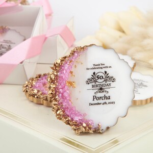 Puede incluir: Favores de fiesta en forma de geoda rosa y dorada con un centro blanco. Los favores tienen un borde dorado y est&aacute;n decorados con purpurina rosa. El centro del favor tiene una inscripci&oacute;n negra que dice "Gracias por celebrar con nosotros. 50 cumplea&ntilde;os Porcha 4 de diciembre de 2023."