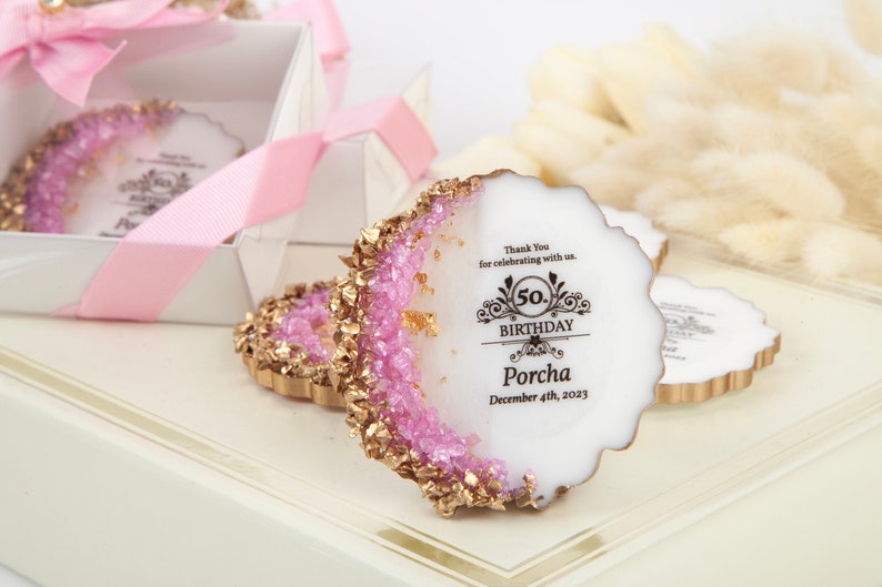 Puede incluir: Favores de fiesta en forma de geoda rosa y dorada con un centro blanco. Los favores tienen un borde dorado y est&aacute;n decorados con purpurina rosa. El centro del favor tiene una inscripci&oacute;n negra que dice "Gracias por celebrar con nosotros. 50 cumplea&ntilde;os Porcha 4 de diciembre de 2023."