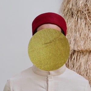 Pode incluir: Uma pessoa usando um fez bordô e uma camisa creme com gola mandarim. A camisa tem uma carcela abotoada. O fundo inclui uma parede texturizada de cor natural.