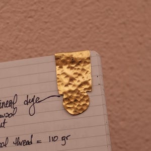 May include: A gold-colored metal bookmark with a hammered, textured surface. The bookmark has a rectangular top section that transitions into a rounded bottom shape. It is clipped onto the corner of a page with handwritten text in black ink.