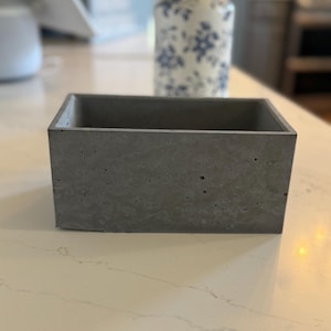 May include: A rectangular, gray concrete planter. The planter has a simple, modern design with a smooth, textured surface. The open top allows for planting small succulents or other decorative items. The planter is suitable for indoor or outdoor use.