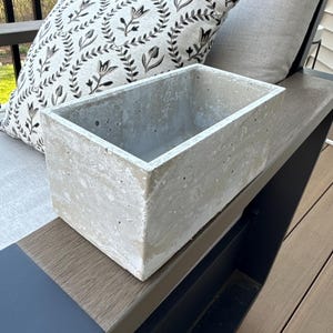 May include: A rectangular concrete planter with a weathered, light gray finish. The planter sits on a wooden surface next to a decorative pillow with a black and white floral pattern. The pillow rests against a gray cushion.