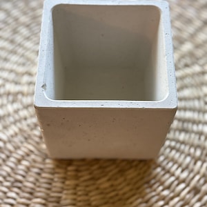 May include: A square, light gray concrete planter with a textured surface. The planter has a rectangular opening and is set against a woven, light brown background. The planter is empty.