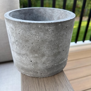 May include: A cylindrical, gray concrete planter with a rough, textured surface. The planter is a light gray color and has a simple, modern design. The planter is suitable for indoor or outdoor use.
