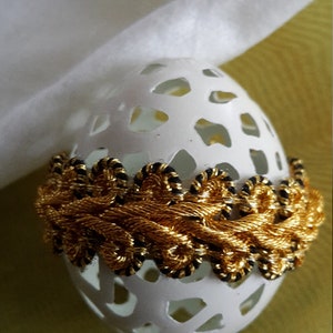 May include: A white egg with a delicate lace design is decorated with a gold and black trim. The egg is sitting on a green surface.
