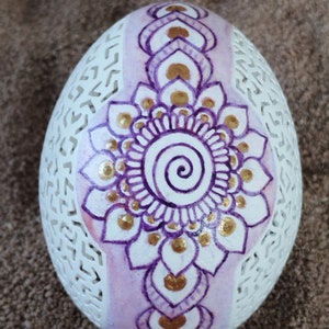 May include: A white egg with a purple and gold mandala design. The egg is decorated with a delicate pattern of flowers and swirls.