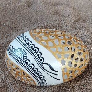 May include: A white egg with a gold and black design. The egg has a geometric pattern on one side and a floral design on the other side.