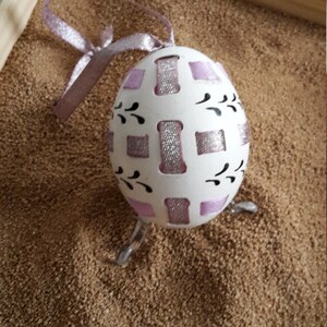 May include: A white egg with pink and silver ribbon designs and black floral accents is displayed on a stand. The egg is decorated with a geometric pattern.
