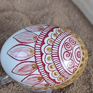 May include: A white egg decorated with red and gold paint. The egg has a mandala design with swirls and dots. The egg is sitting on a stand.