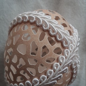 May include: A decorative, hollow eggshell with a peach-colored hue, featuring a delicate, openwork design. The egg is adorned with a white, embroidered trim that adds a touch of elegance. This handcrafted item is ideal for Easter or home decor.