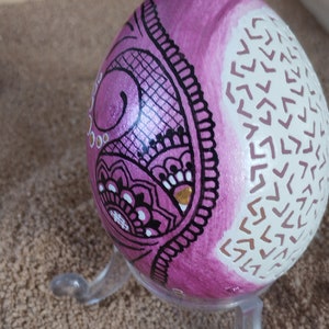 May include: A pink egg with a black and white mandala design on one side and a white background with a white chevron pattern on the other side. The egg is on a clear plastic stand.
