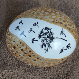 May include: A white egg with a delicate gold lattice design and a black silhouette of a dandelion with seeds blowing in the wind. The egg is sitting on a brown surface.