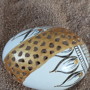 May include: A white egg with a gold and black design. The egg has a geometric pattern cut out of the gold paint, revealing the white egg shell underneath. The design includes black floral shapes and gold accents.