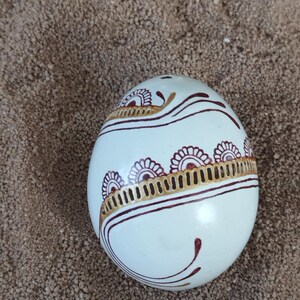May include: A white egg with intricate red and gold designs. The egg is decorated with a swirling pattern of lines and dots.