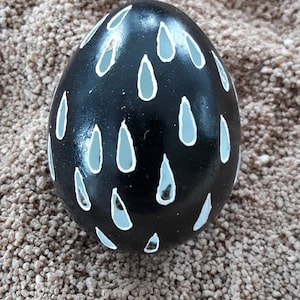 May include: A black Easter egg with white raindrop designs painted on it. The egg is sitting on a bed of brown sand.