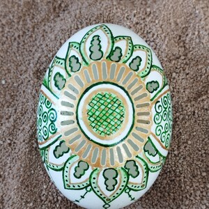 May include: A white egg with a green and gold mandala design. The egg is decorated with intricate patterns and swirls.