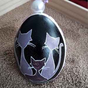 May include: A black egg-shaped ornament with three purple cats painted on it. The cats are in a sitting position and have white whiskers. The ornament has a white pearl on top.