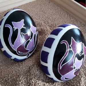 May include: Two painted decorative stones with a black background and a white border. Each stone features a purple cat design. The stones are displayed on a bed of sand inside a wooden tray.