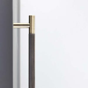 May include: A vertical door pull with a textured, dark gray center grip and brass-colored ends. The handle is mounted on a white surface, with a gray edge visible on the left side.