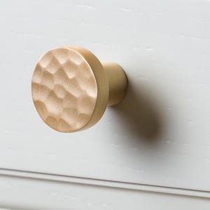 May include: Close-up of a gold cabinet knob with a textured, hammered surface. The round knob is mounted on a light gray cabinet door with a recessed panel. A wooden countertop is visible in the background.