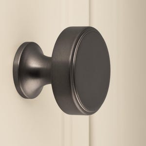 Puede incluir: Un pomo de armario redondo gris oscuro con una cara plana y una base circular más pequeña. El pomo está montado en una puerta de color crema. El herraje tiene un acabado mate y un diseño sencillo y moderno.