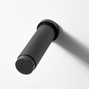 Puede incluir: Tres objetos cilíndricos negros con extremos redondeados están dispuestos diagonalmente sobre un fondo blanco. Dos tienen una superficie texturizada y estriada, mientras que el del medio es liso. Los objetos proyectan largas sombras.