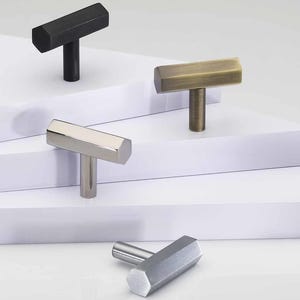 May include: A collection of cabinet knobs in various metallic finishes, including black, brass, and silver. Each knob features a T-shaped design with a hexagonal bar. The Heritage Brass logo is visible in the bottom right corner.