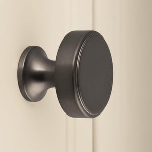 May include: A dark bronze cabinet knob with a circular face and a cylindrical base is mounted on a cream-colored door. The knob has a simple, modern design, and the door has vertical paneling.