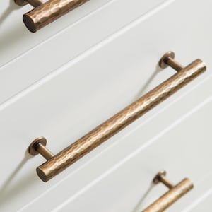 May include: Four white cabinet drawers with brushed gold cabinet pulls. The pulls are hammered and have a slightly rounded rectangular shape.