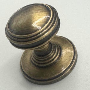 May include: A bronze-colored cabinet knob with a rounded top and a circular base. The knob features a series of concentric rings around the top and base, adding a decorative element. The hardware is designed for use on cabinets and drawers.