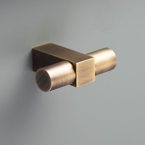 Puede incluir: Un tirador de armario en tono latón con base rectangular y asa cilíndrica. El herraje está montado sobre una superficie gris claro, con un borde blanco visible en la esquina superior izquierda. Diseño moderno y minimalista.