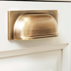 May include: A close-up of a bronze-colored cabinet pull mounted on a white cabinet door. The pull has a rectangular backplate and a curved, cup-like handle. The hardware has a brushed finish.