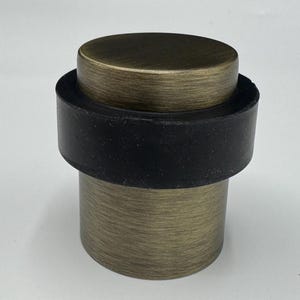 May include: A bronze-colored door stop with a black rubber ring. The door stop has a cylindrical shape with a flat top and a textured finish. The product is designed to prevent doors from slamming shut.