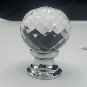 May include: A decorative crystal ball finial with a faceted surface, mounted on a shiny silver-colored base. The finial is designed to be used as a decorative element for curtains or other home decor items.