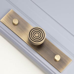 Op de afbeelding: Een messing kastgreep met een cirkelvormig ontwerp. De greep heeft een rechthoekige achterplaat met twee kleine, ronde accenten. De hoofdknoppunt heeft concentrische cirkels. De hardware is gemonteerd op een lichtgrijze ondergrond.