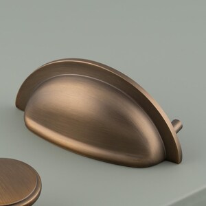 May include: A set of three cabinet hardware pieces in a brushed bronze finish, including one semi-circular cup pull and two round knobs. Each piece features a smooth, metallic surface with subtle dark highlights and a rounded, classic profile.