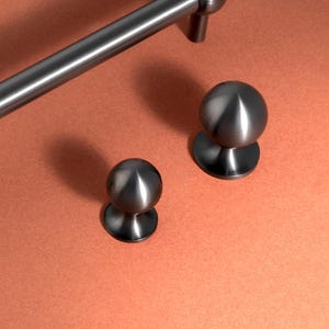 May include: A collection of brushed nickel cabinet hardware, featuring two bar pulls and two round knobs. The pulls have a cylindrical bar with rounded ends. The knobs have a spherical shape. The hardware is displayed on a copper-coloured surface.