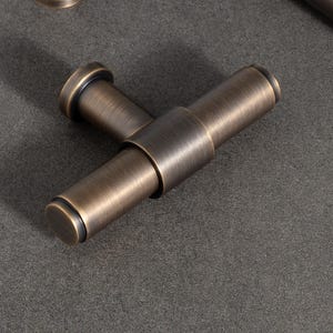 Puede incluir: Juego de herrajes para armarios en tono bronce, que incluye un tirador, un pomo y un tirador en forma de T. El herraje tiene un diseño cilíndrico con extremos redondeados y un acabado mate. Las piezas están dispuestas sobre una superficie gris.