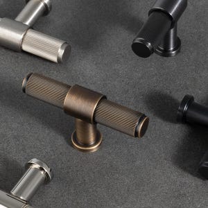 May include: A collection of six T-bar cabinet knobs in various metallic finishes including brushed gold, matte black, polished silver, and antique bronze. Each knob features a cylindrical handle with a textured, ribbed grip and a central post.